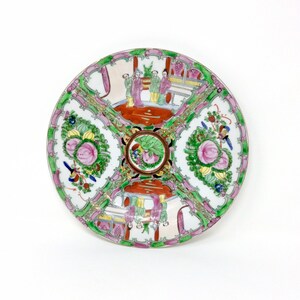 May include: A white porcelain plate with a green, pink, and gold floral design. The center of the plate features a floral motif with a green and gold border. The rim of the plate is decorated with a pink and green floral design.