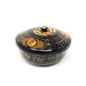 May include: A black lacquered trinket box with a lid. The box is decorated with gold and orange geometric patterns and a yin yang symbol.