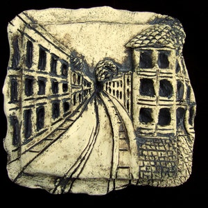 May include: A ceramic relief sculpture depicting a street scene with buildings on either side of a narrow street. The buildings are rendered in a simple, stylized manner, with black lines outlining the windows and doors. The street is paved with cobblestones, and there is a tree in the center of the scene.