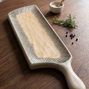 Puede incluir: Una tabla de servir rectangular de madera con un centro texturizado de color claro y un borde oscuro tallado y acanalado. La tabla cuenta con un mango redondeado en un extremo. La superficie tiene un acabado de veta de madera natural.