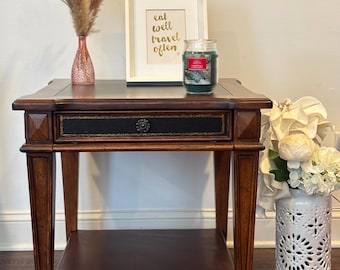 May include: A dark wood side table with a drawer and a lower shelf. The table has decorative legs and small wheels. A framed print with the text "eat well travel often" sits on top, along with a candle and a vase with dried flowers.