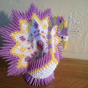 Könnte beinhalten: Ein lila, weißer und gelber Origami-Pfau mit einer weißen Perlenkrone. Der Pfau steht auf einer Holzoberfläche.