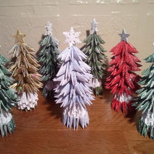 Può includere: Sette alberi di Natale origami in vari colori, tra cui verde, oro, bianco e rosso. Ogni albero ha una stella o un fiocco di neve in cima ed è decorato con glitter.