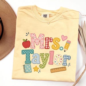 May include: A pale yellow t-shirt featuring the text Mrs. Taylor in colorful, patterned block letters. The design includes a red apple, a yellow pencil, a small notebook, a star, a ruler, and hearts, all rendered in a patchwork style graphic.
