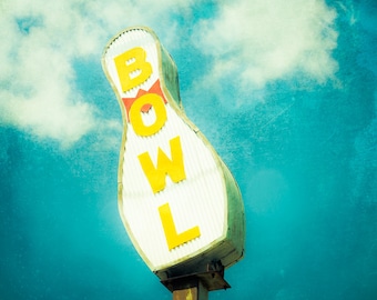 May include: A white bowling pin shaped sign with yellow letters spelling "BOWL" against a blue sky with white clouds.