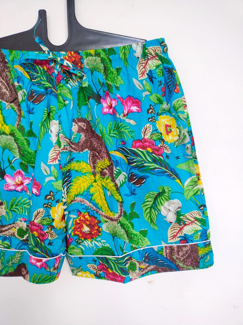 May include: Turquoise shorts with a jungle-themed print, including monkeys, flowers, and foliage. The shorts have a drawstring waist and white trim. The fabric is a bright turquoise colour.