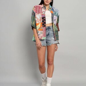 Op de afbeelding: Een overhemd met korte mouwen en knoopsluiting met een veelkleurig patchworkontwerp met diverse bloemen- en geometrische patronen, gedragen over een witte crop top met een lichtblauwe spijkerbroekshort, witte sokken en witte sportschoenen.