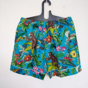 May include: Turquoise shorts with a vibrant floral and monkey print. The shorts have a white trim and are hanging on a black hanger. The design includes pink, yellow, and green foliage.