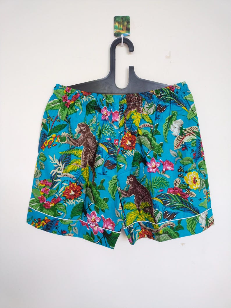May include: Turquoise shorts with a vibrant floral and monkey print. The shorts have a white trim and are hanging on a black hanger. The design includes pink, yellow, and green foliage.
