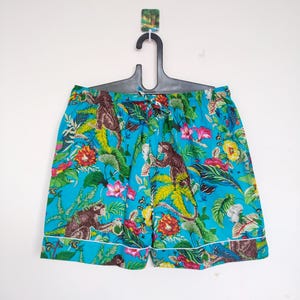 May include: Turquoise shorts with a jungle print featuring monkeys, flowers, and foliage. The shorts have a white trim and are hanging on a black hanger.