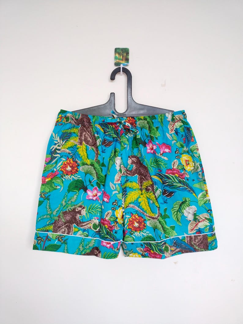 May include: Turquoise shorts with a jungle print featuring monkeys, flowers, and foliage. The shorts have a white trim and are hanging on a black hanger.