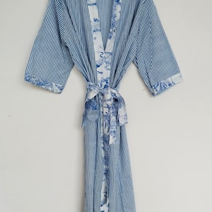 Könnte beinhalten: Blau-weiß gestreifter Bademantel mit passendem Bindegürtel. Der Bademantel hat eine kontrastierende weiße Bordüre mit blauem Blumendesign an den Kanten und am Bindegürtel. Der Bademantel hängt an einem schwarzen Kleiderbügel.