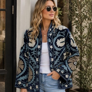 Può includere: Una giacca blu navy aperta sul davanti con intricati ricami floreali in azzurro e beige. Il capo presenta maniche lunghe, colletto alla coreana e orlo dritto, indossato sopra una canotta bianca e jeans in denim di colore azzurro.