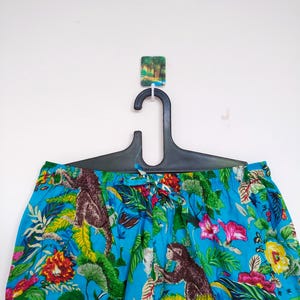 May include: Blue patterned trousers with a jungle theme, featuring brown monkeys, pink and yellow flowers, and lush green foliage. The trousers are hanging on a black hanger against a white background.