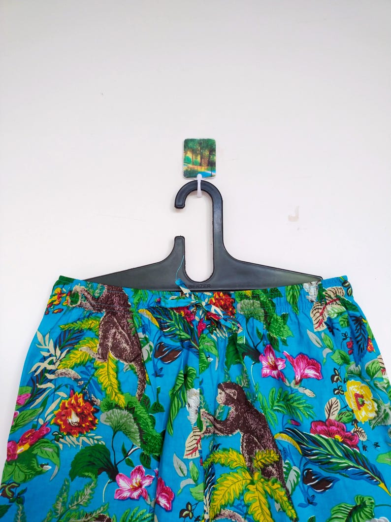 May include: Blue patterned trousers with a jungle theme, featuring brown monkeys, pink and yellow flowers, and lush green foliage. The trousers are hanging on a black hanger against a white background.