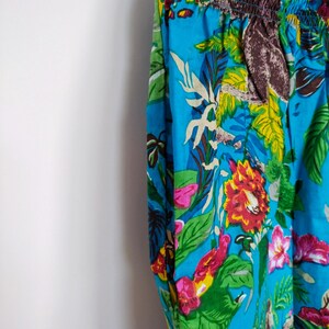 May include: Close-up of blue patterned trousers. The fabric is decorated with a colourful floral design, including red, pink, and yellow flowers, green leaves, and brown animal figures. The waistband is elasticated.