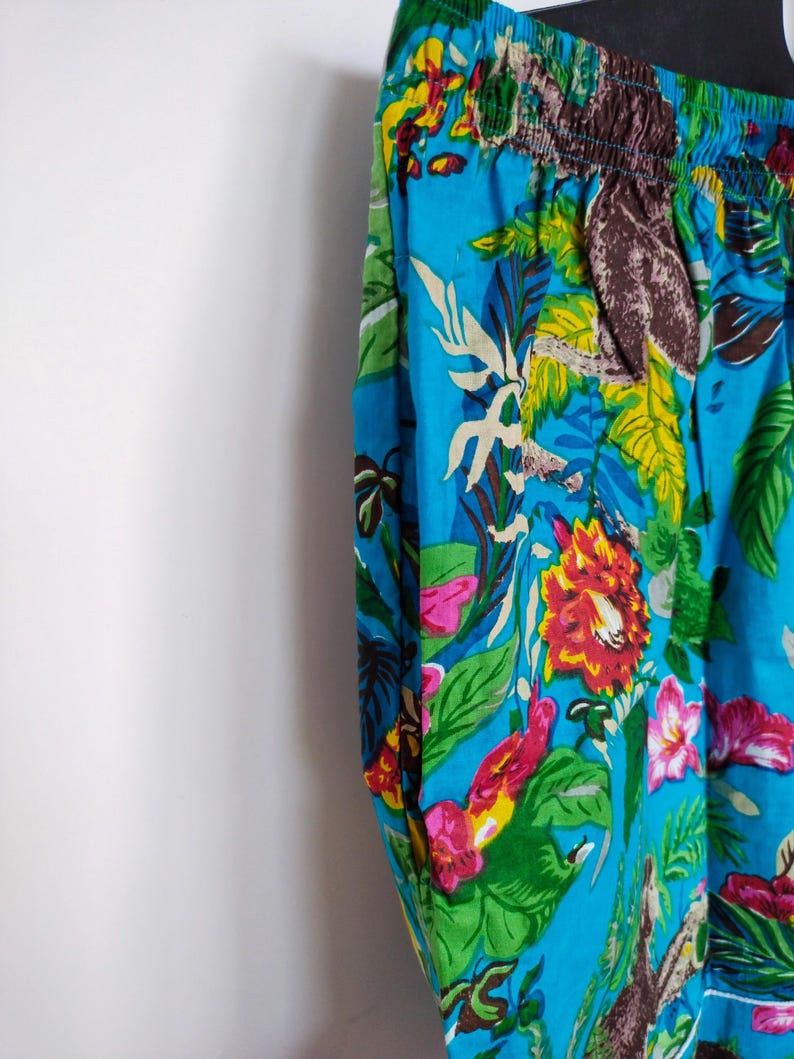 May include: Close-up of blue patterned trousers. The fabric is decorated with a colourful floral design, including red, pink, and yellow flowers, green leaves, and brown animal figures. The waistband is elasticated.