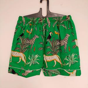 May include: Green shorts with a tropical print featuring zebras, leopards, toucans, and palm trees. The shorts have a white trim and an elastic waistband. The fabric is a vibrant green color.