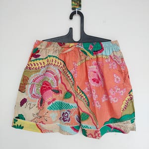 May include: Floral print shorts in shades of peach, pink, green, and beige. The shorts feature a drawstring waist and white trim along the hem. The fabric is patterned with large, stylized flowers and leaves. The shorts are hanging on a black hanger.
