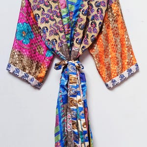 May include: A colorful, patterned kimono robe with long sleeves. The robe features a mix of floral, paisley, and geometric designs in shades of pink, blue, orange, and tan. It is tied at the waist with a matching sash and hangs from a wooden hanger.