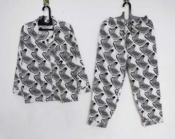 May include: A long-sleeve button-down pajama shirt and matching drawstring pajama pants featuring a repeating black and white zebra head pattern. The set is displayed on black plastic hangers against a plain white background.