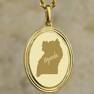 Könnte beinhalten: Goldfarbene Halskette mit Anhänger. Der runde Anhänger zeigt den Umriss von Uganda und den Namen des Landes in einer Schreibschrift. Der Anhänger hängt an einer passenden goldenen Seilkette. Der Hintergrund ist eine neutrale, strukturierte Farbe.
