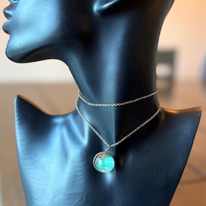 May include: A silver necklace with a double chain and a round pendant. The pendant features a colourful, abstract design with shades of teal, orange, and red. The necklace is displayed on a black mannequin.