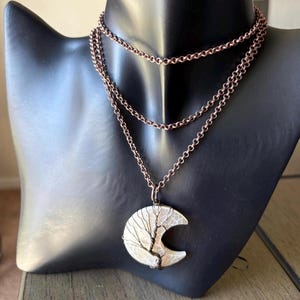 May include: A copper-colored necklace with three strands of chain. The pendant is a crescent moon shape with a wire tree design. The necklace is displayed on a black mannequin.