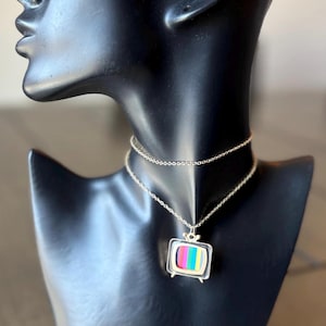 May include: A silver-toned necklace featuring a pendant shaped like a retro television set. The TV screen displays colorful vertical stripes. The necklace is displayed on a black mannequin.