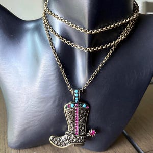 May include: A gold-toned necklace featuring a pendant shaped like a cowboy boot. The boot is adorned with turquoise and pink gemstones, and intricate scrollwork. The necklace has a three-strand chain.