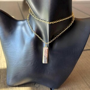 May include: A gold-toned double-layered chain necklace featuring a rectangular pendant with a marbled, multi-colored stone inlay. The necklace consists of a shorter upper chain and a longer lower chain that holds the vertical rectangular charm.