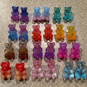 Peut inclure: Une collection de boucles d'oreilles en forme d'oursons en gélatine colorés, dans diverses nuances de bleu, violet, rouge, orange et vert. Les boucles d'oreilles sont disposées par paires, mettant en valeur leur aspect vibrant et translucide. Ces boucles d'oreilles fantaisie sont un accessoire amusant.