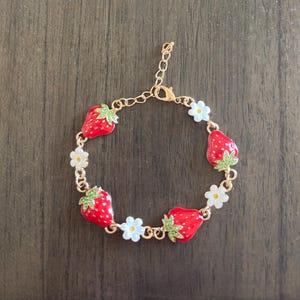 Peut inclure: Un bracelet doré orné de breloques fraises rouges avec des feuilles vertes et des fleurs blanches. Le bracelet est réglable et possède une fermeture à fermoir homard. Un bijou charmant.