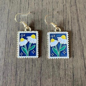 May include: A pair of gold-tone hook earrings featuring rectangular pendants shaped like postage stamps. The pendants have a dark blue background with white daisies, green stems, yellow centers, and small star accents, plus the text 4¢.