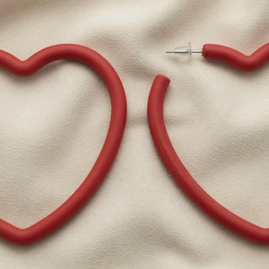 May include: A pair of large, matte red heart-shaped hoop earrings. Each earring features a smooth, rounded frame in the shape of a heart with a thin metal post and a clear plastic backing for secure attachment to the earlobe.