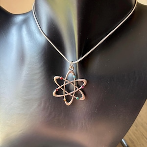 May include: Silver-toned necklace featuring a pendant shaped like an atom, with colorful gemstone accents. The pendant is suspended from a delicate chain. The necklace is displayed on a black mannequin.