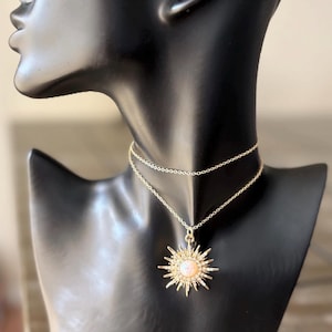 May include: A gold-colored necklace with a sunburst pendant, displayed on a black mannequin. The pendant features a central, light-colored stone surrounded by radiating spikes. The necklace has a delicate chain.