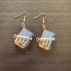 May include: A pair of gold-tone drop earrings featuring a rectangular blue enamel sardine tin charm with the text SARDINES visible. Each charm includes a small metal key attached to the front and depicts three silver fish inside the tin.