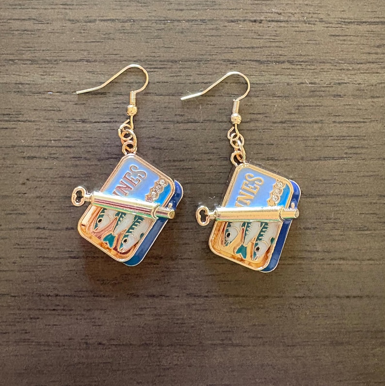 May include: A pair of gold-tone drop earrings featuring a rectangular blue enamel sardine tin charm with the text SARDINES visible. Each charm includes a small metal key attached to the front and depicts three silver fish inside the tin.