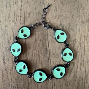 May include: A black metal bracelet featuring eight light green alien head charms. Each charm has black oval eyes and a black outline. The bracelet is connected by small black links and has a lobster clasp closure.