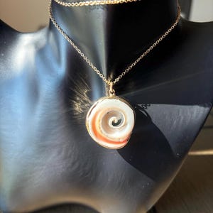 Puede incluir: Un collar dorado con un colgante con diseño de espiral. El colgante tiene un esquema de color blanco y naranja, enmarcado en dorado. El collar se exhibe en un maniquí negro.