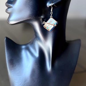 May include: A silver-toned dangle earring featuring a miniature rectangular sardine tin charm with blue and white enamel details, a small metal key attached to the front, and the partial text "NES" visible on the top surface of the tin.