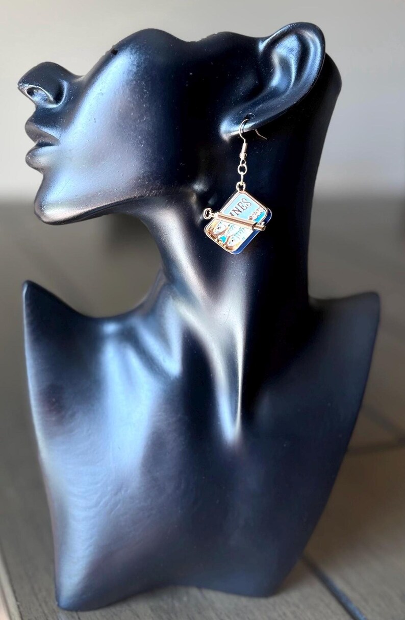 May include: A silver-toned dangle earring featuring a miniature rectangular sardine tin charm with blue and white enamel details, a small metal key attached to the front, and the partial text "NES" visible on the top surface of the tin.