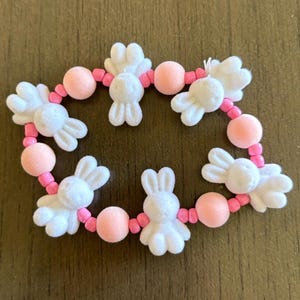 May include: A bracelet featuring white bunny charms and pink beads. The bunny charms have a soft, fuzzy texture, and the beads are a solid, light pink. The bracelet is strung on a pink cord, creating a playful and festive accessory.