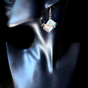 May include: A dangling earring featuring a miniature rectangular tin of sardines with a metallic key attached to the side. The label on the tin is blue and white with the partial text NES visible. The earring hangs from a thin gold-toned hook.