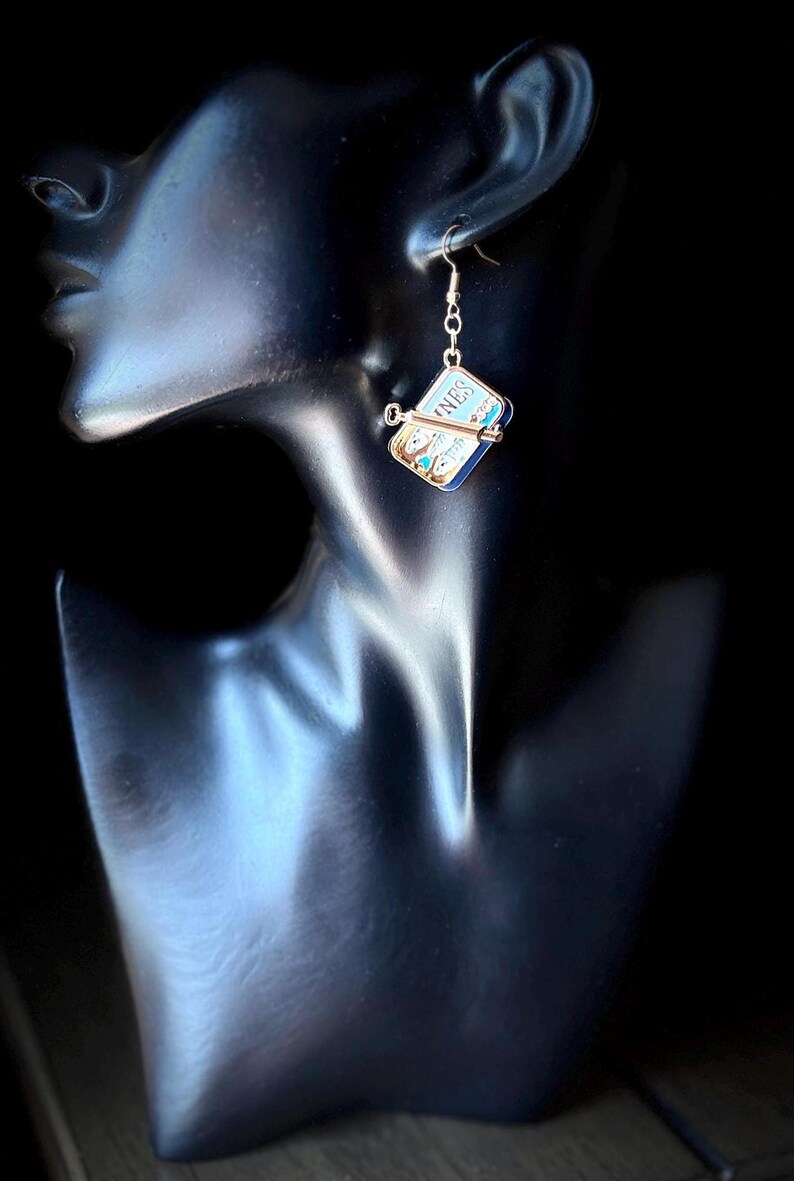 May include: A dangling earring featuring a miniature rectangular tin of sardines with a metallic key attached to the side. The label on the tin is blue and white with the partial text NES visible. The earring hangs from a thin gold-toned hook.