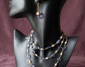 May include: A multi-strand necklace and a single earring set featuring small white oval pearls and translucent purple oval beads linked by gold-toned metal chain. The necklace drapes in three layers, while the earring hangs from a gold hook.