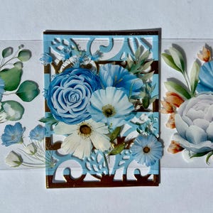 Pode incluir: Design floral decorativo com flores azuis e brancas, incluindo rosas e margaridas, dispostas dentro de uma moldura azul clara com detalhes dourados. O design está sobre uma superfície transparente, com elementos florais adicionais.