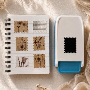 Puede incluir: Una perforadora de papel blanca y azul con una abertura rectangular en forma de sello, colocada junto a un cuaderno de espiral que contiene seis pegatinas de papel marrón con ilustraciones de flores y patrones de cuadrícula en negro.