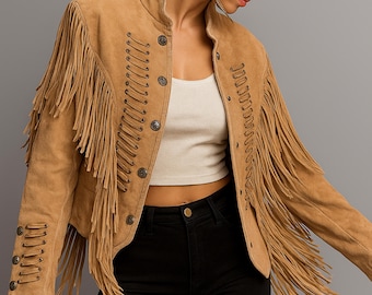 May include: A tan suede jacket with long fringe detailing. The jacket features a round collar, decorative stitching, and button closures. The sleeves and front panels are adorned with long fringe. The jacket is paired with black jeans.